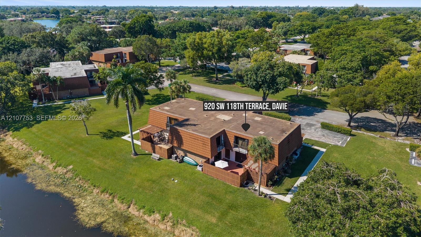 Multi-level exterior featuring a shingled mansard roofline, private ground-level patio, balcony, and waterfront positioning on a manicured lot with mature palm trees