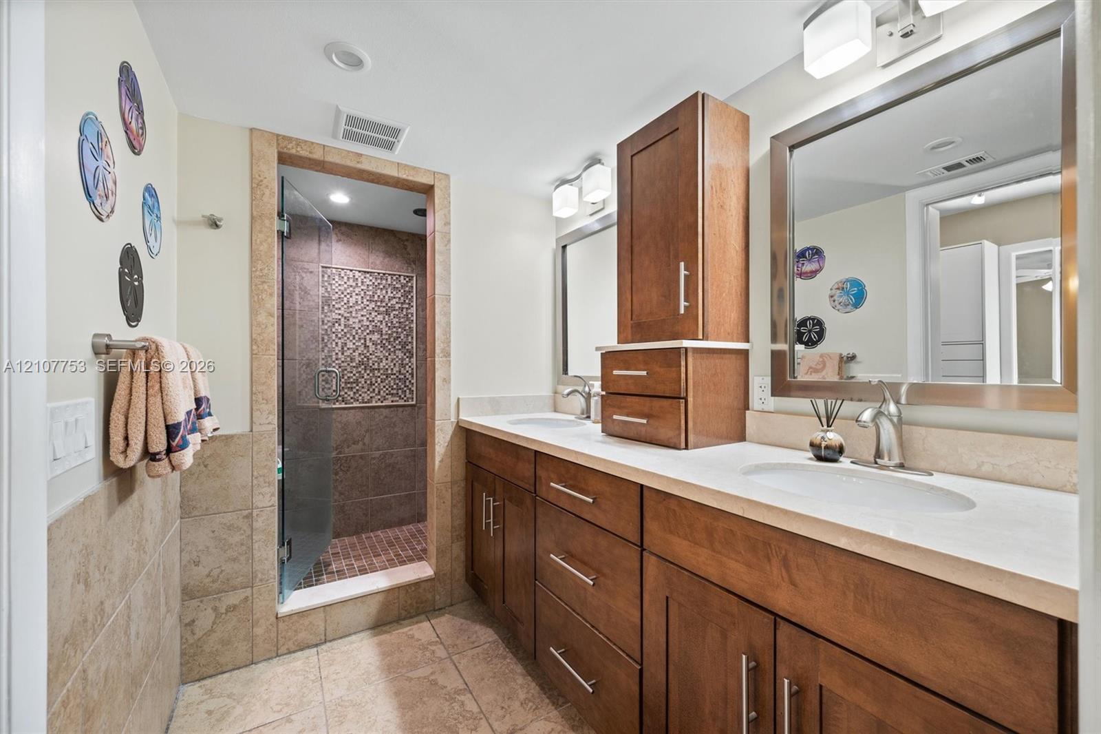 Double vanity featuring wood-finish cabinetry, solid-surface countertops, dual rectangular under-mount sinks, and a glass-enclosed shower with tiled walls and decorative mosaic inlay