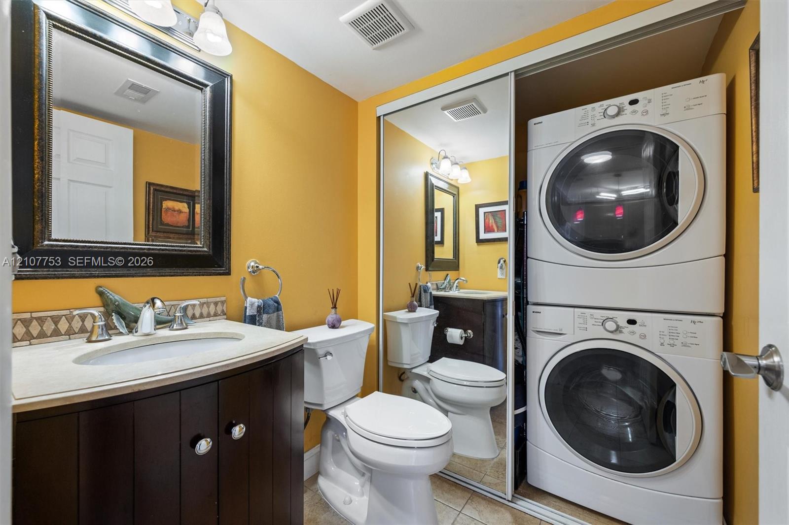 Bathroom featuring dark wood-finish cabinetry, a neutral countertop with an integrated basin, mirrored sliding closet doors, and stacked laundry appliances