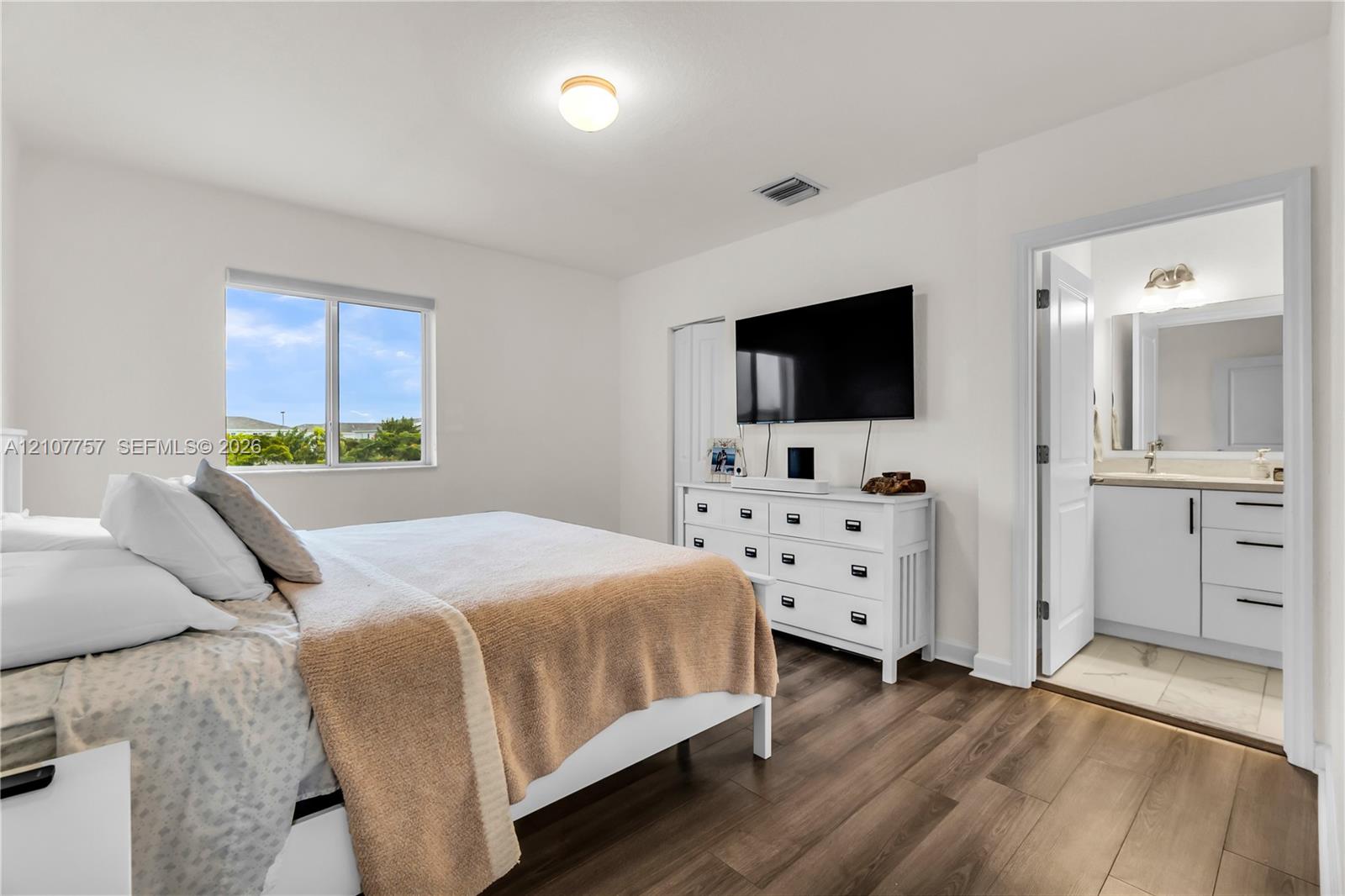 Wood-finish flooring, neutral walls, large window with natural light, and an ensuite bathroom featuring a vanity with drawers