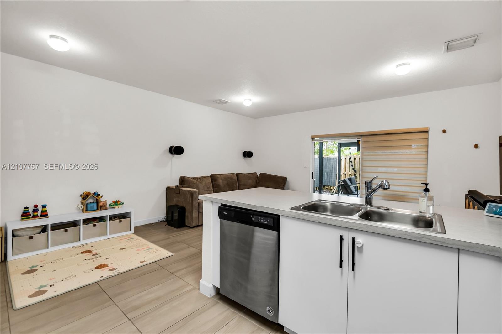 Kitchen island featuring a stainless steel dishwasher, dual-basin stainless steel sink with a high-arch faucet, white cabinetry with matte black hardware, and wood-finish tile flooring