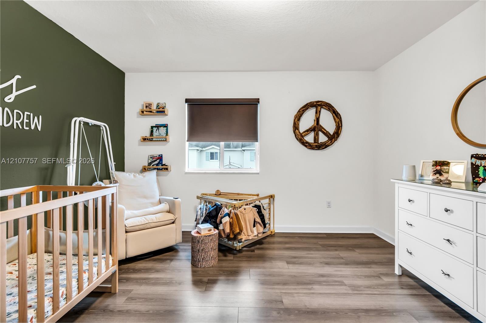 Room featuring dark wood-finish flooring, a dark green accent wall, a dual-pane window with a roller shade, and three wall-mounted floating shelves