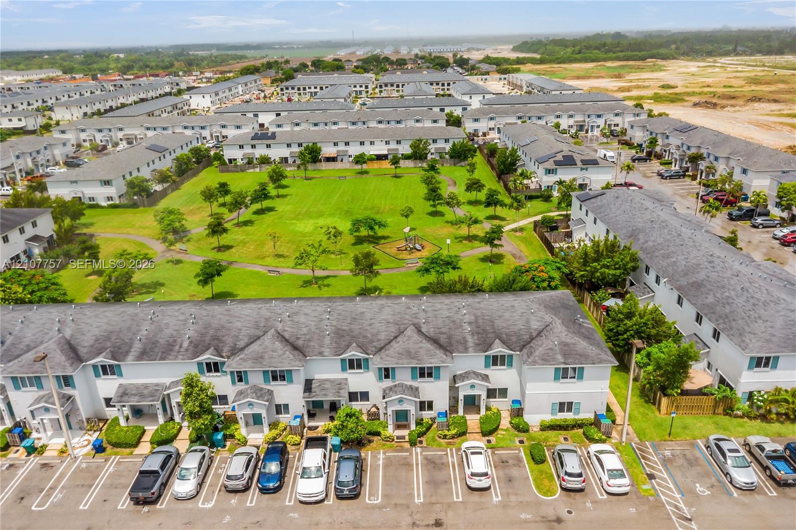 Aerial perspective of a multi-unit residential complex featuring light-toned siding, dark shingle roofs, designated surface parking, and extensive landscaped green space with paved pedestrian walkways