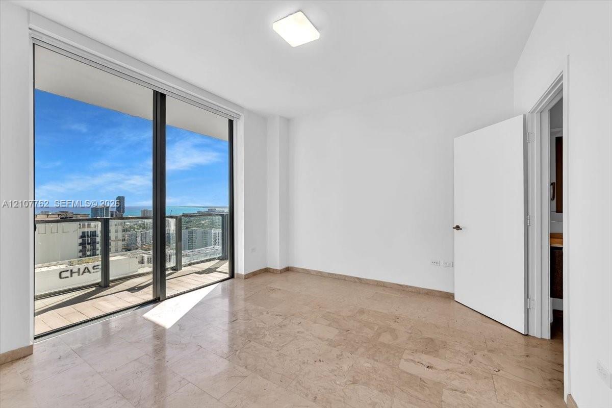 Floor-to-ceiling sliding glass doors, private balcony, neutral stone tile flooring, and overhead flush-mount lighting