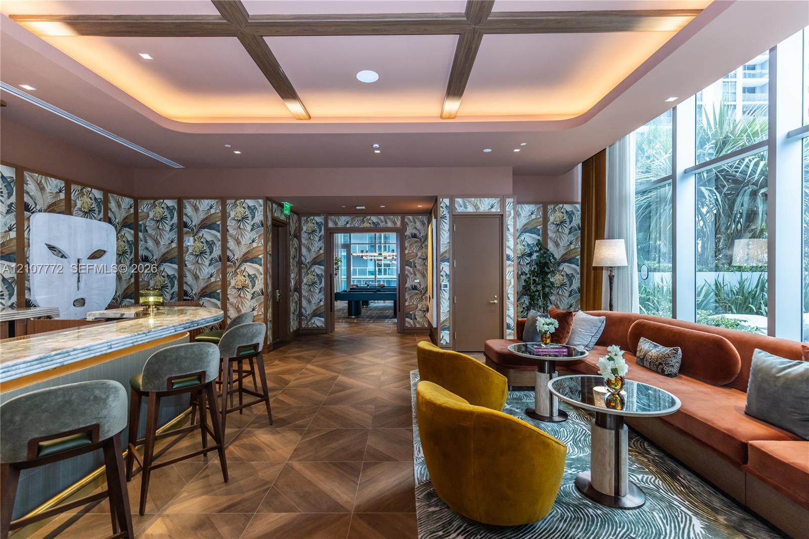 Coffered ceiling with integrated cove lighting, wood-finish flooring in a chevron pattern, floor-to-ceiling windows, built-in bar, and custom wall treatments