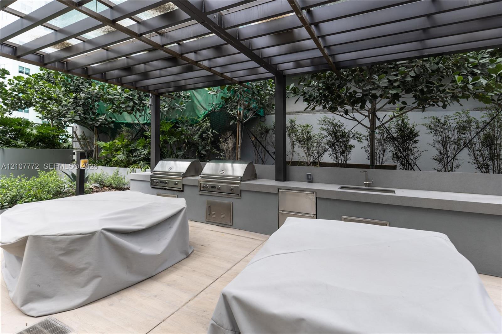 Outdoor kitchen featuring dual stainless steel grills, integrated stainless steel storage, a built-in sink, and a permanent pergola structure set over concrete hardscaping