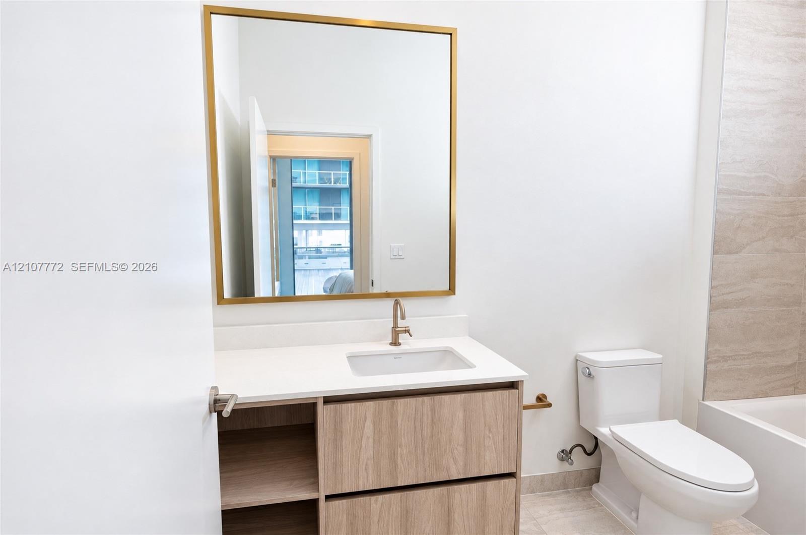 Bathroom featuring a vanity with wood-finish cabinetry, white countertop with an integrated basin, gold-toned hardware, and a square wall mirror with a metallic frame Bathroom featuring a vanity with wood-finish cabinetry, white countertop with an integrated basin, gold-toned hardware, and a square wall mirror with a metallic frame