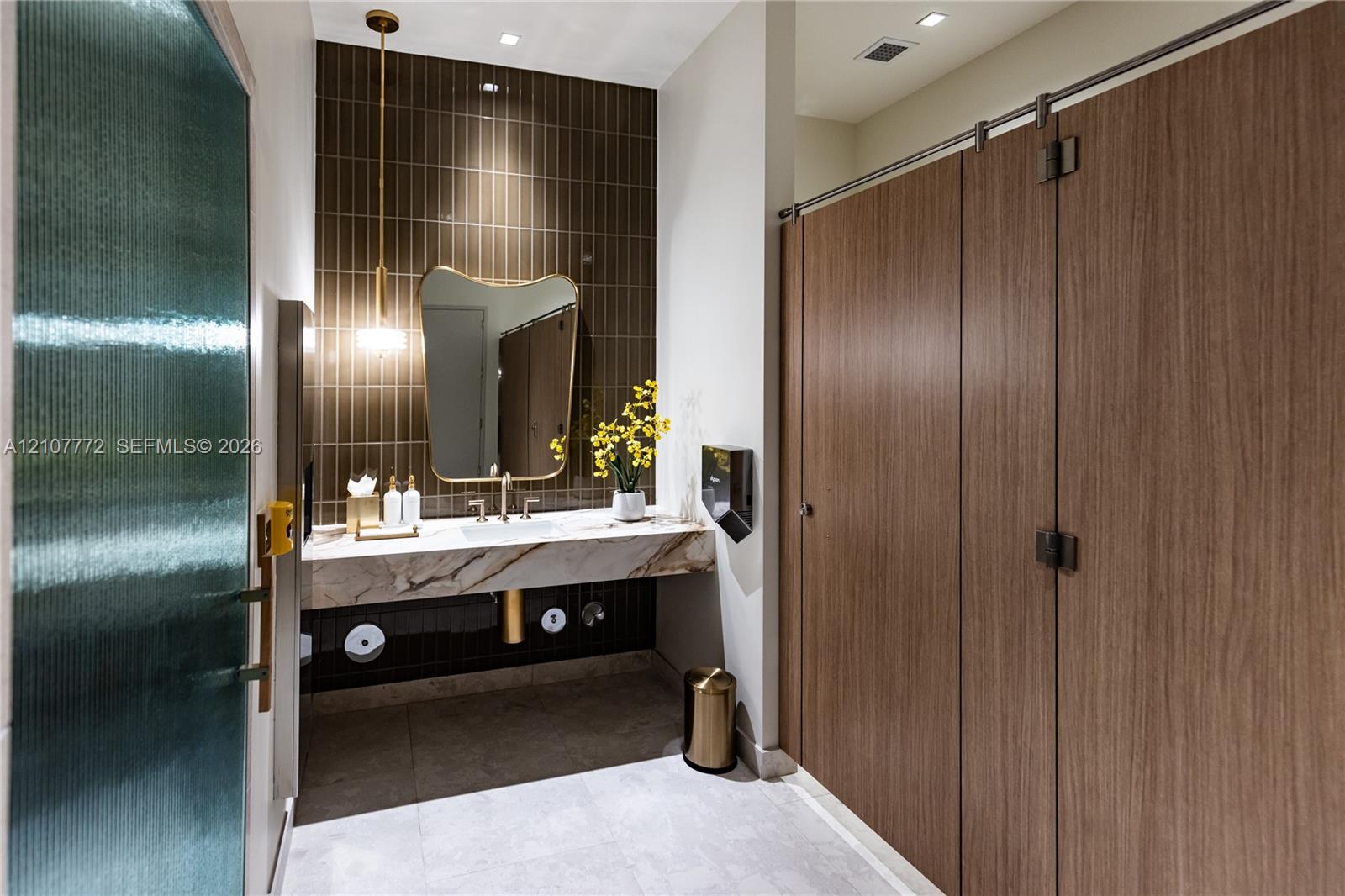 Floating vanity with a stone countertop, vertical tile backsplash, gold-tone pendant lighting, irregular-shaped mirror, and wood-finish partition doors