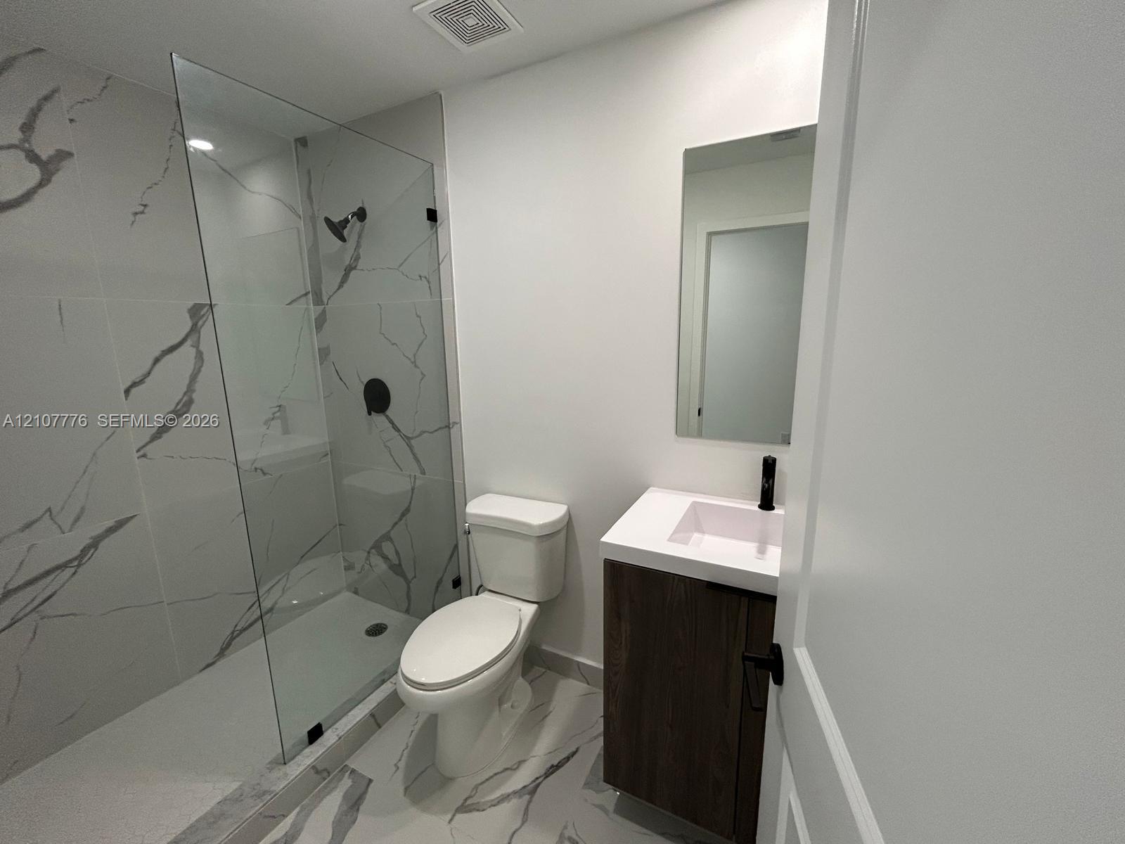 Walk-in shower with floor-to-ceiling veined tile, wood-finish floating vanity with a white countertop, matte black fixtures, and a frameless glass enclosure