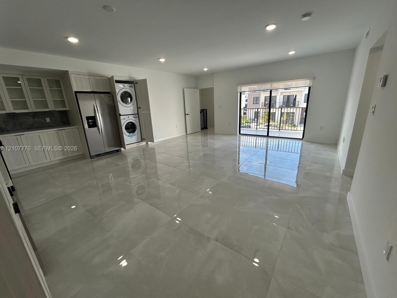 Open-concept interior featuring large-format tile flooring, recessed lighting, white cabinetry with glass-front uppers, stainless steel refrigerator, and a sliding glass door providing external access