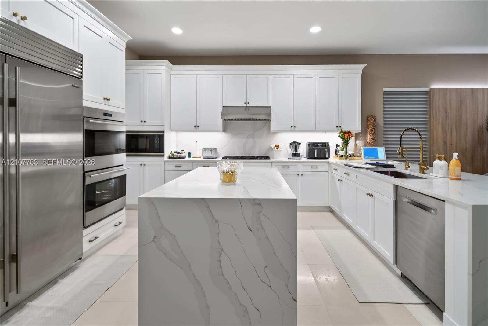 Kitchen featuring white shaker cabinetry, a waterfall-edge center island with stone-finish countertops, stainless steel appliances, a professional-style range hood, and gold-tone faucet hardware