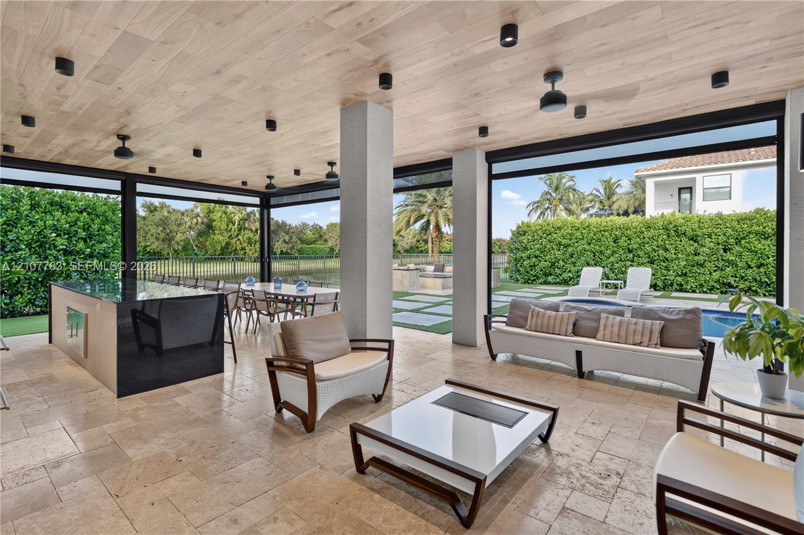 Covered outdoor living space featuring natural stone flooring, wood-plank ceiling, recessed lighting, and a built-in outdoor kitchen with a dark-toned island