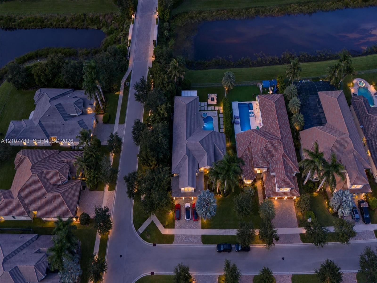 Aerial perspective of residential properties featuring tile roofing, paved driveways, private swimming pools, and landscaped perimeters situated along a waterfront boundary