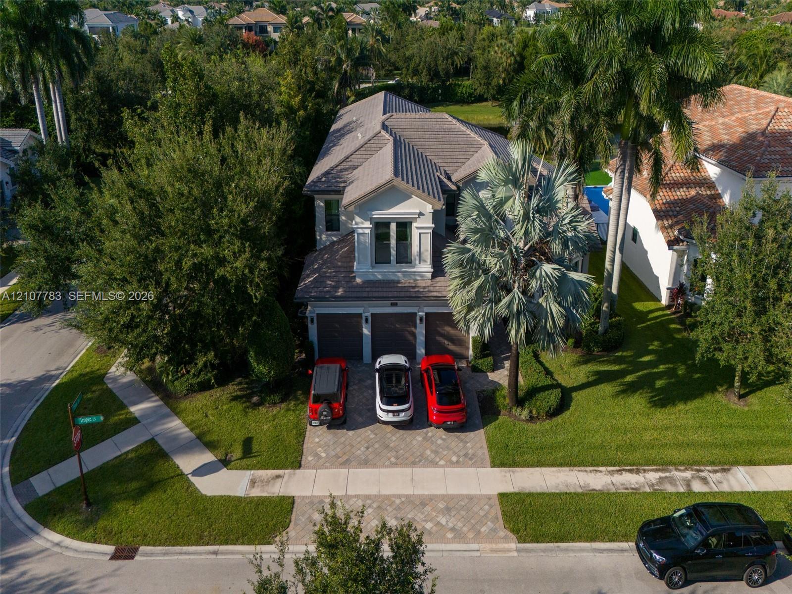 Aerial perspective of a multi-story residence featuring a tile roof, three-car garage, and a paved stone driveway situated on a landscaped lot with mature palm trees
