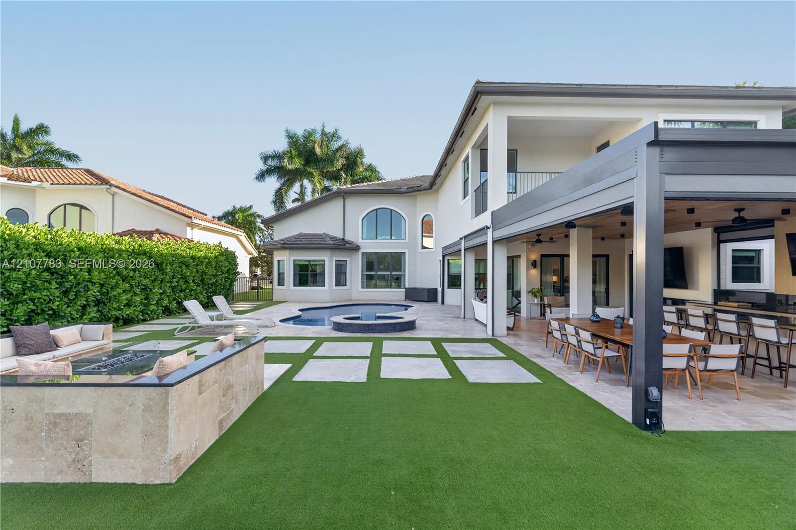 Outdoor living area featuring a covered pergola, stone-clad fire pit, swimming pool with attached spa, and artificial turf lawn with stone pavers