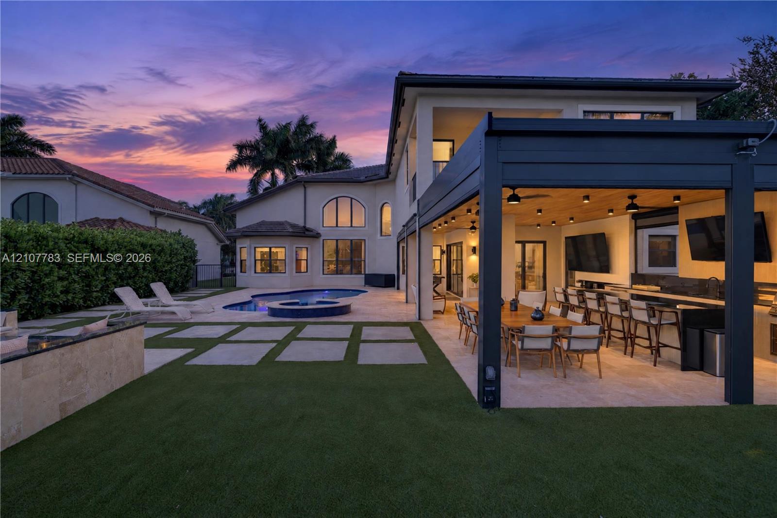 Rear exterior featuring a covered patio with a built-in outdoor kitchen, a swimming pool with an integrated spa, stone-paver hardscaping, and synthetic turf lawn