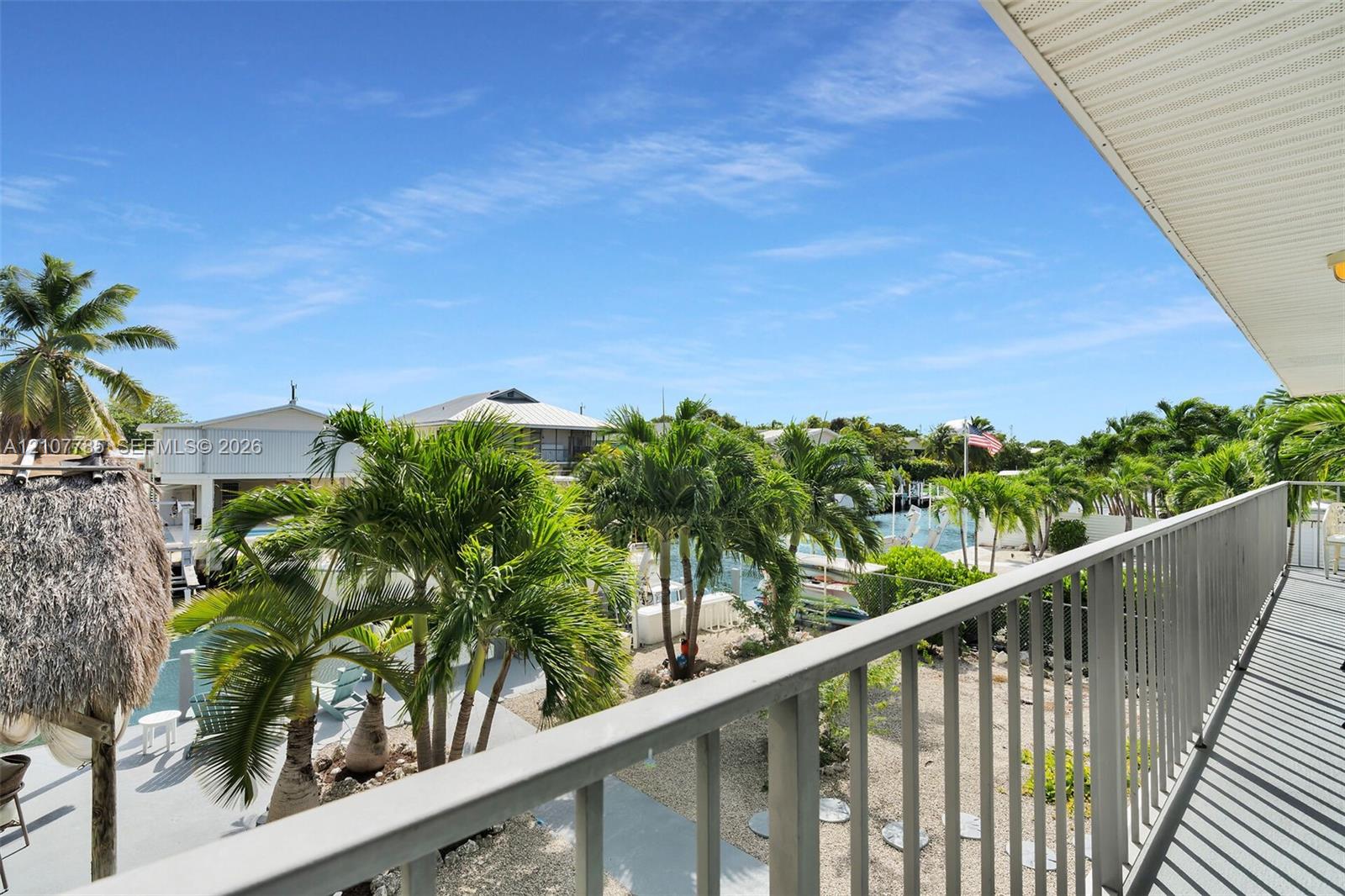 Elevated balcony featuring a metal railing and a covered ceiling, overlooking a canal-side yard with mature palm trees and natural landscaping