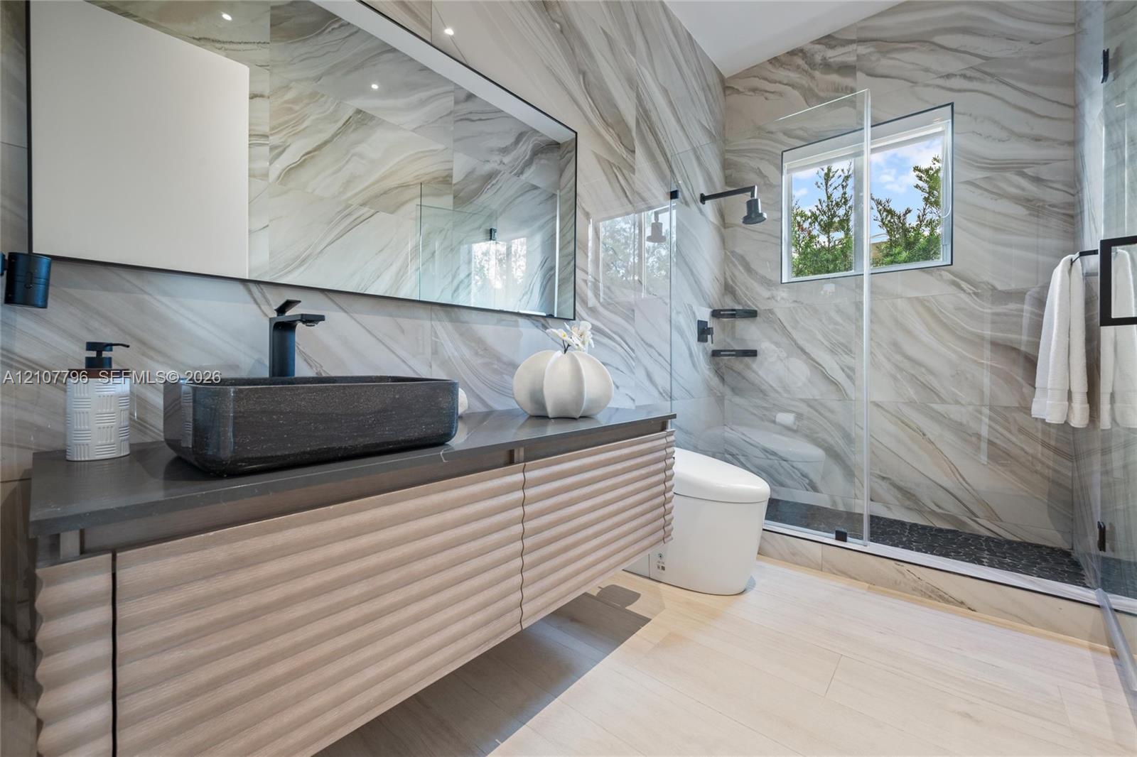 Floating vanity with ribbed cabinetry and solid surface countertop, rectangular vessel sink, wall-spanning mirror, full-height decorative tile wall, and glass-enclosed shower with matte black fixtures