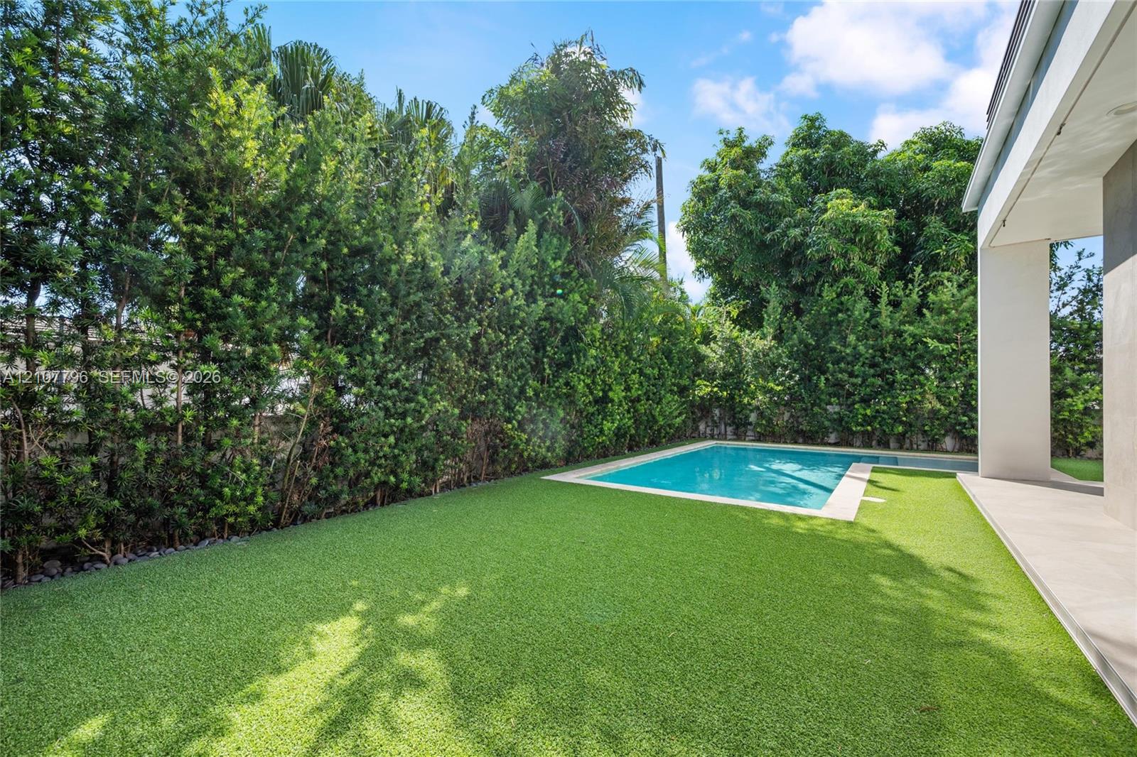 Private backyard featuring an in-ground swimming pool, synthetic turf lawn, stone paver decking, and mature perimeter hedging