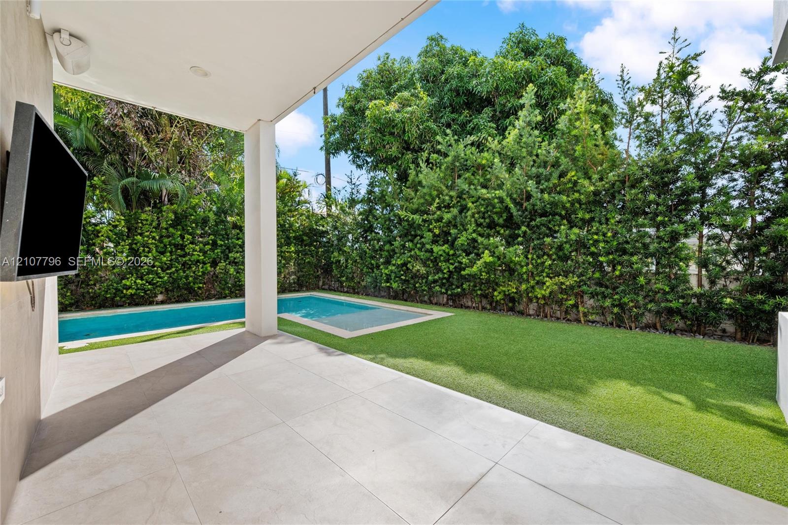 Covered patio with large-format tile flooring, in-ground swimming pool, synthetic turf lawn, and perimeter privacy hedging
