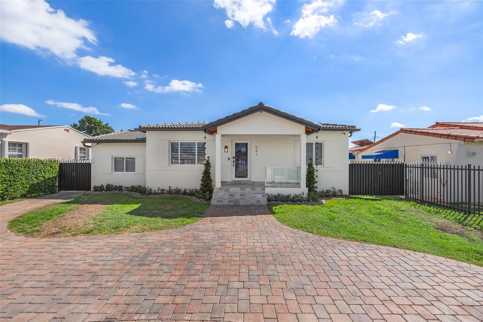 Single-story residence featuring a paved brick driveway, covered entryway with stone steps, glass-panel railing, and metal perimeter fencing