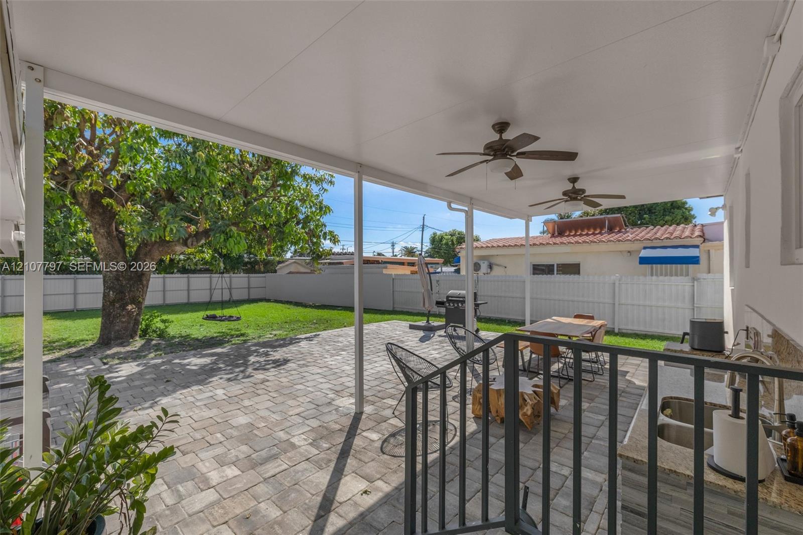 Covered patio featuring a paved stone surface, dual ceiling fans, built-in outdoor kitchen with stone-finish cabinetry, and a vinyl perimeter fence enclosing the lawn