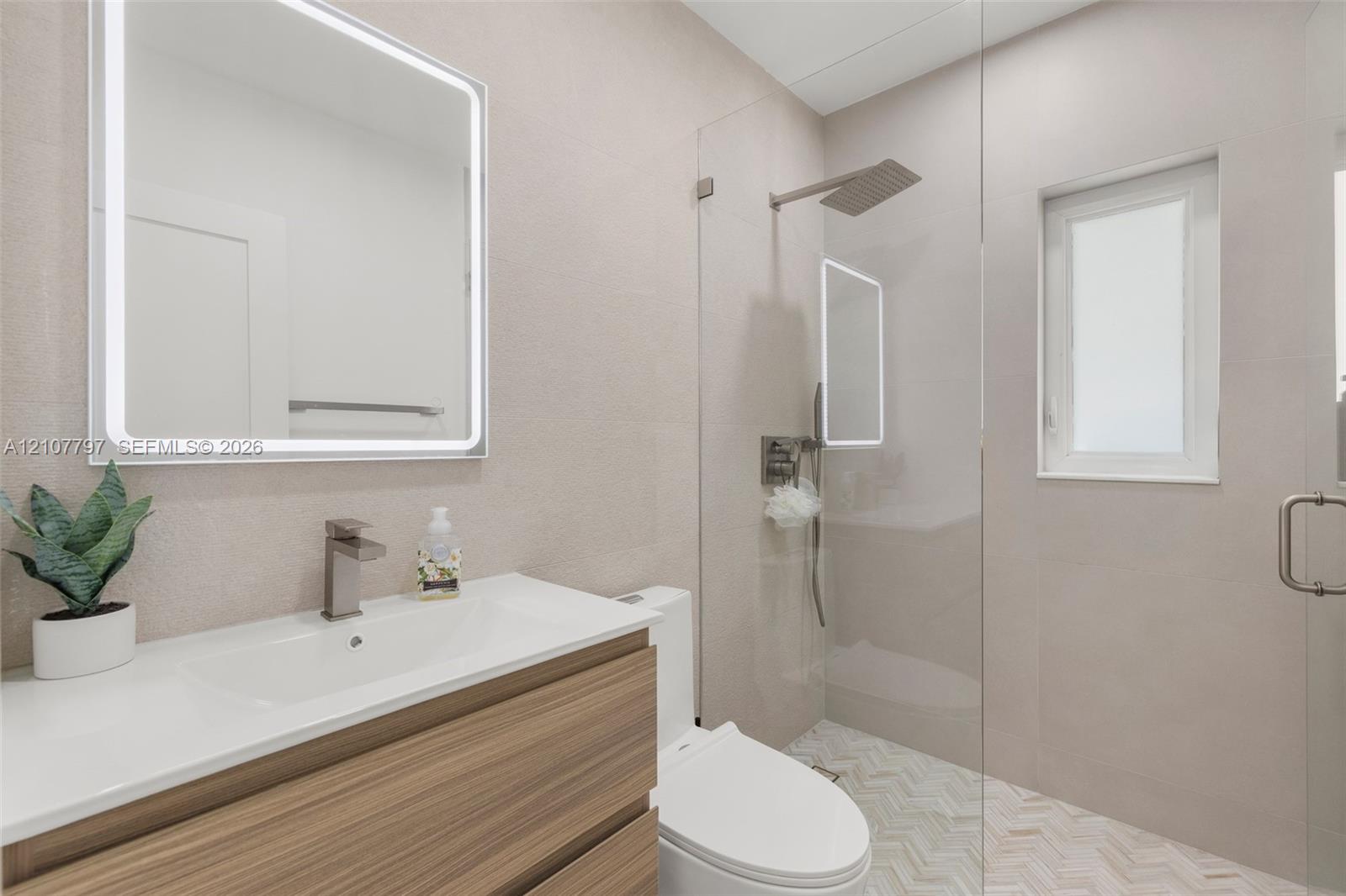 Modern floating vanity with a wood-finish cabinet, integrated sink, wall-mounted illuminated mirror, frameless glass-enclosed shower, and herringbone-patterned floor tile