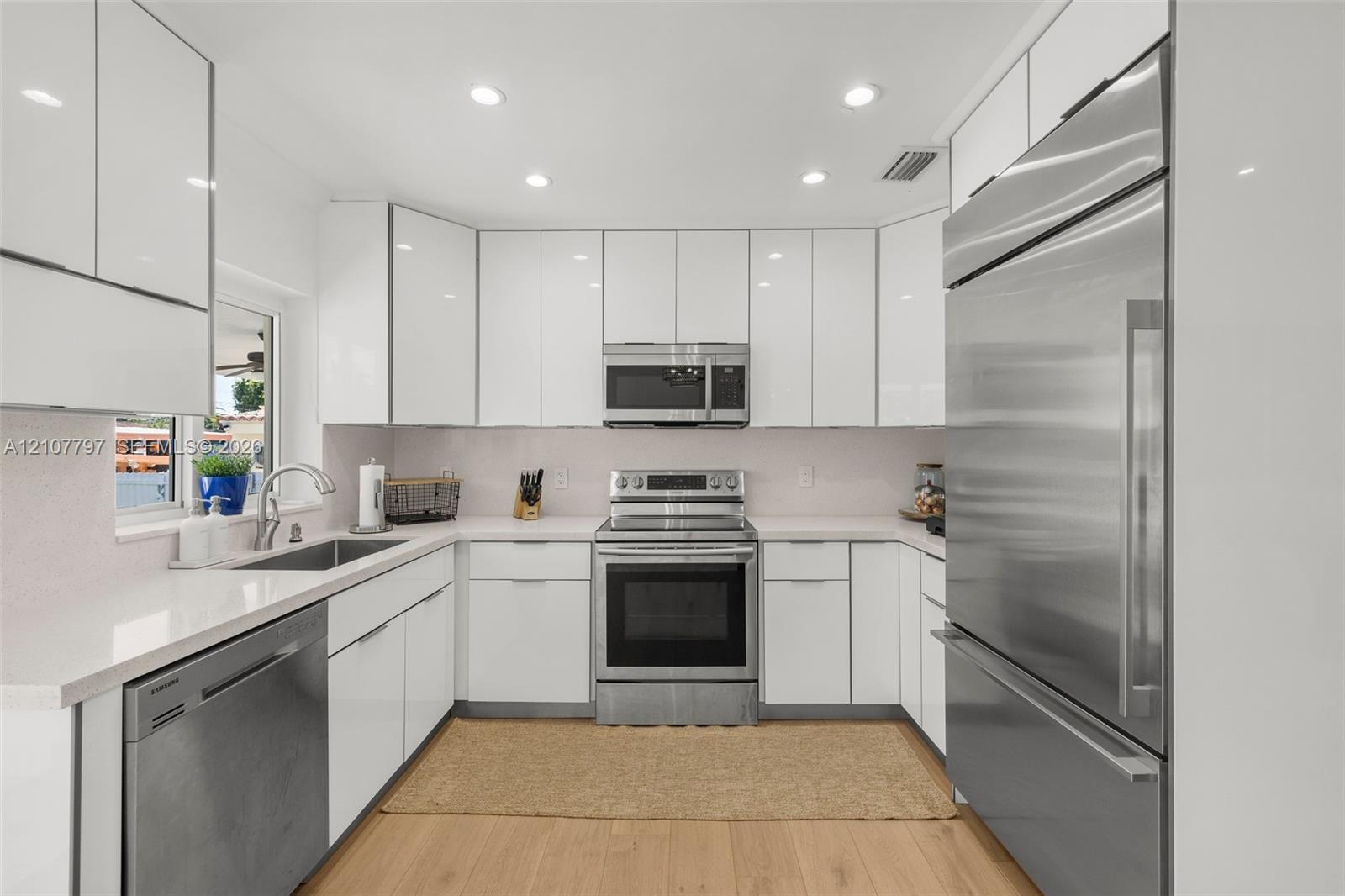 Contemporary kitchen featuring high-gloss white cabinetry, light-toned countertops, wood-finish flooring, and stainless steel appliances including a built-in refrigerator, range, and dishwasher
