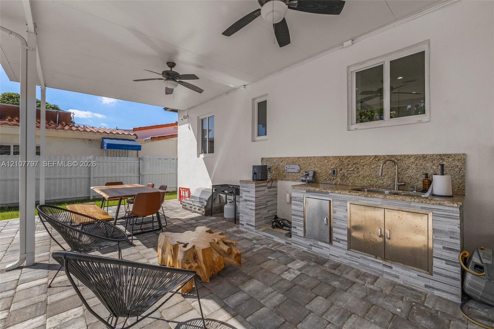 Covered patio featuring paver flooring, two ceiling fans, and an outdoor kitchen with stone veneer base, metal cabinetry, and a stainless steel faucet