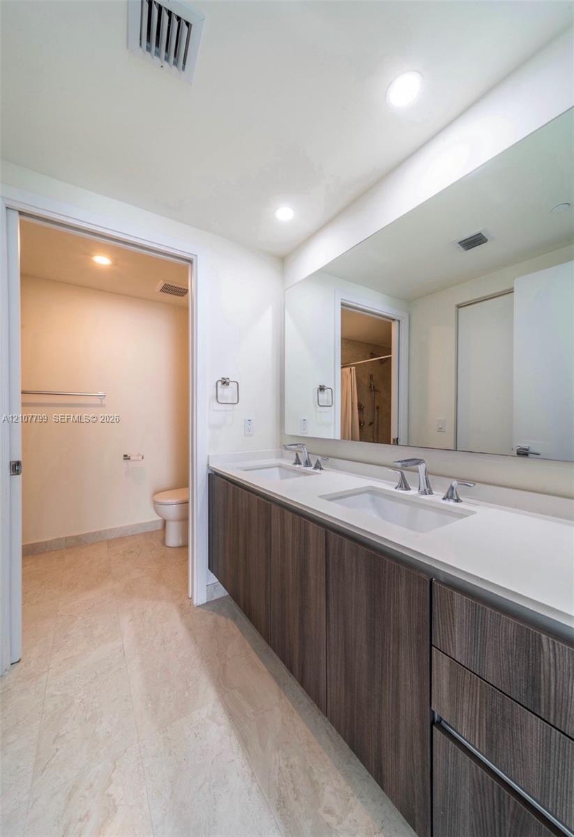 Dual-sink floating vanity with wood-finish cabinetry, solid surface countertop, large wall-mounted mirror, and tile flooring