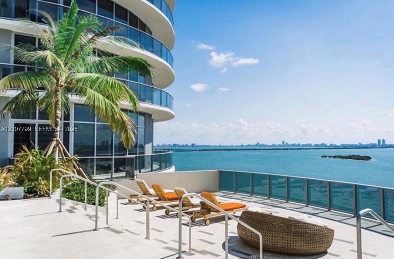 Expansive waterfront terrace featuring light stone tiling, glass panel railings, and metallic handrails