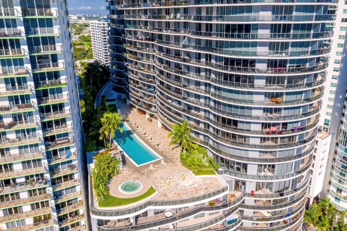 High-rise exterior featuring curved glass balconies, a rectangular swimming pool, a circular spa, tiered hardscaping, and tropical landscaping