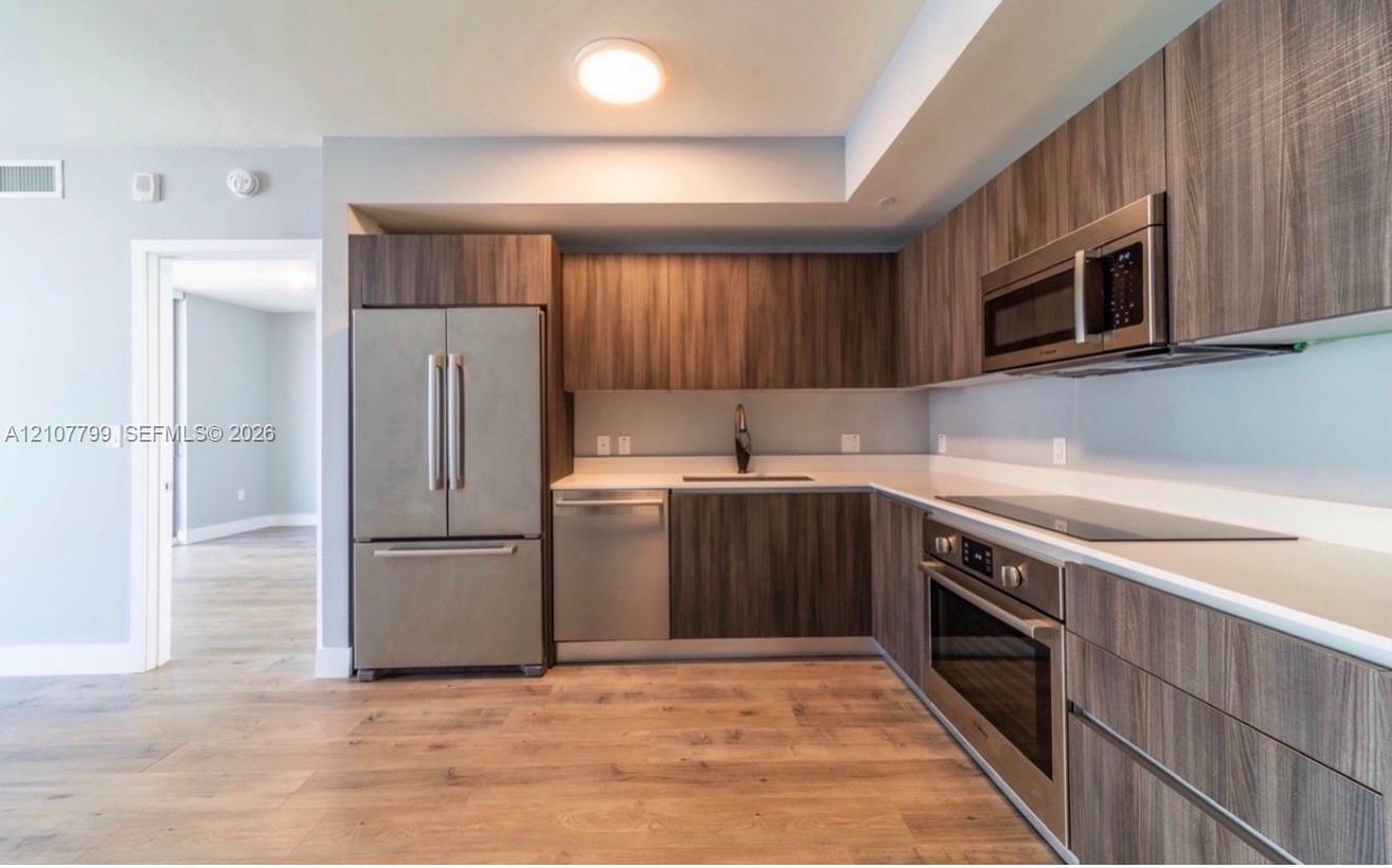 Modern kitchen featuring flat-panel wood-finish cabinetry, stainless steel appliances, solid surface countertops, and wood-finish flooring