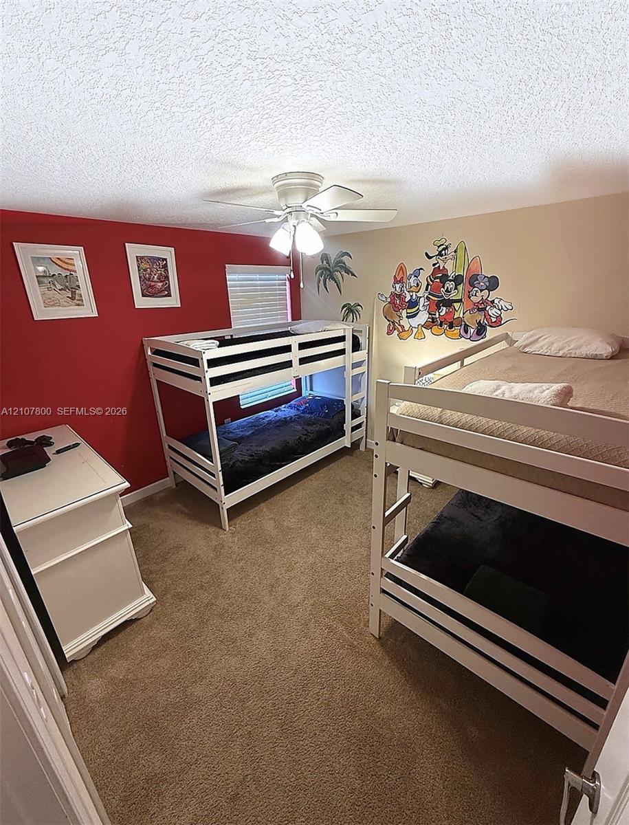 Textured ceiling with a ceiling-mounted light fixture and fan, carpeted flooring, and a single window with horizontal blinds