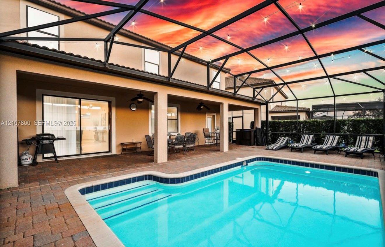 Screened-in pool area featuring paver patio surfacing, integrated ceiling fans, and string lighting against a sunset backdrop