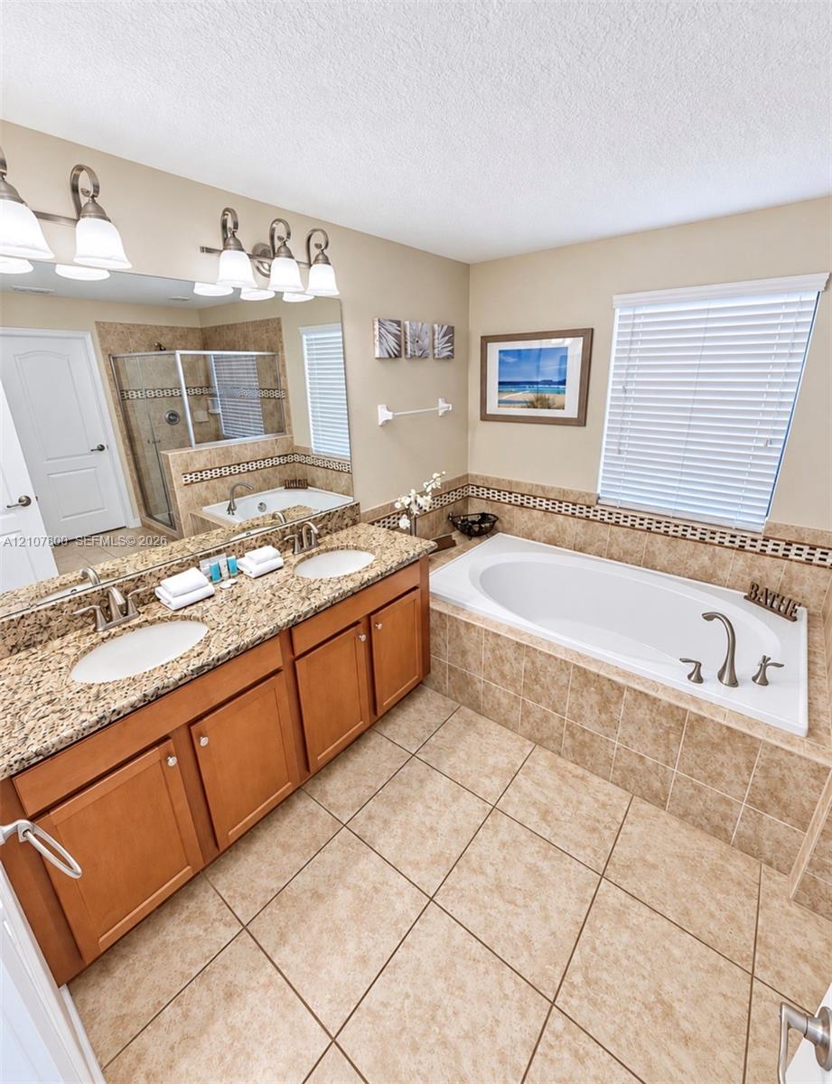 Dual-sink vanity with stone-finish countertop, soaking tub with tile surround, walk-in glass shower, and ceramic tile flooring