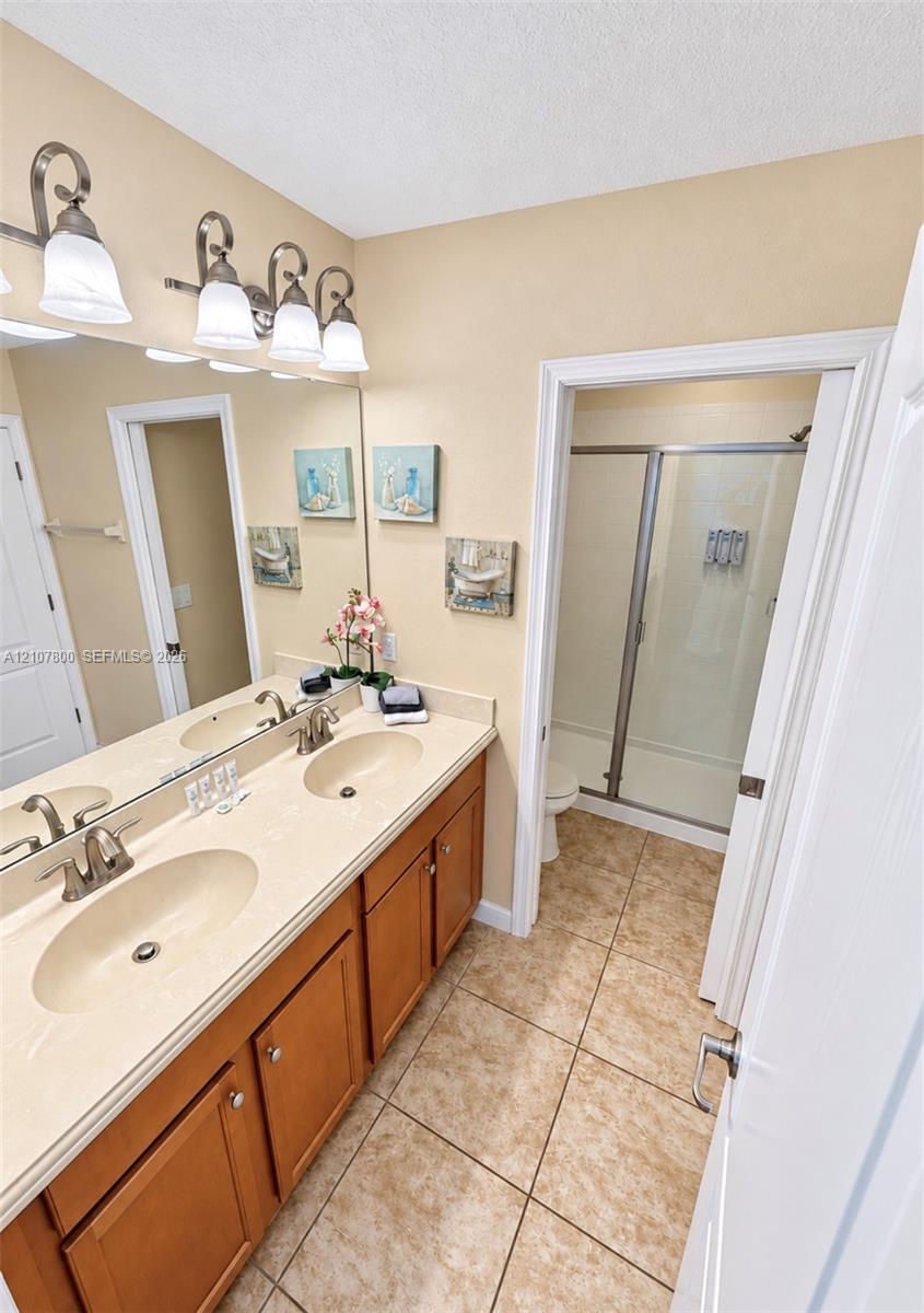 Dual-sink vanity with a solid-surface countertop, four-light vanity fixture, expansive wall-to-wall mirror, tiled flooring, and a walk-in shower with a glass door