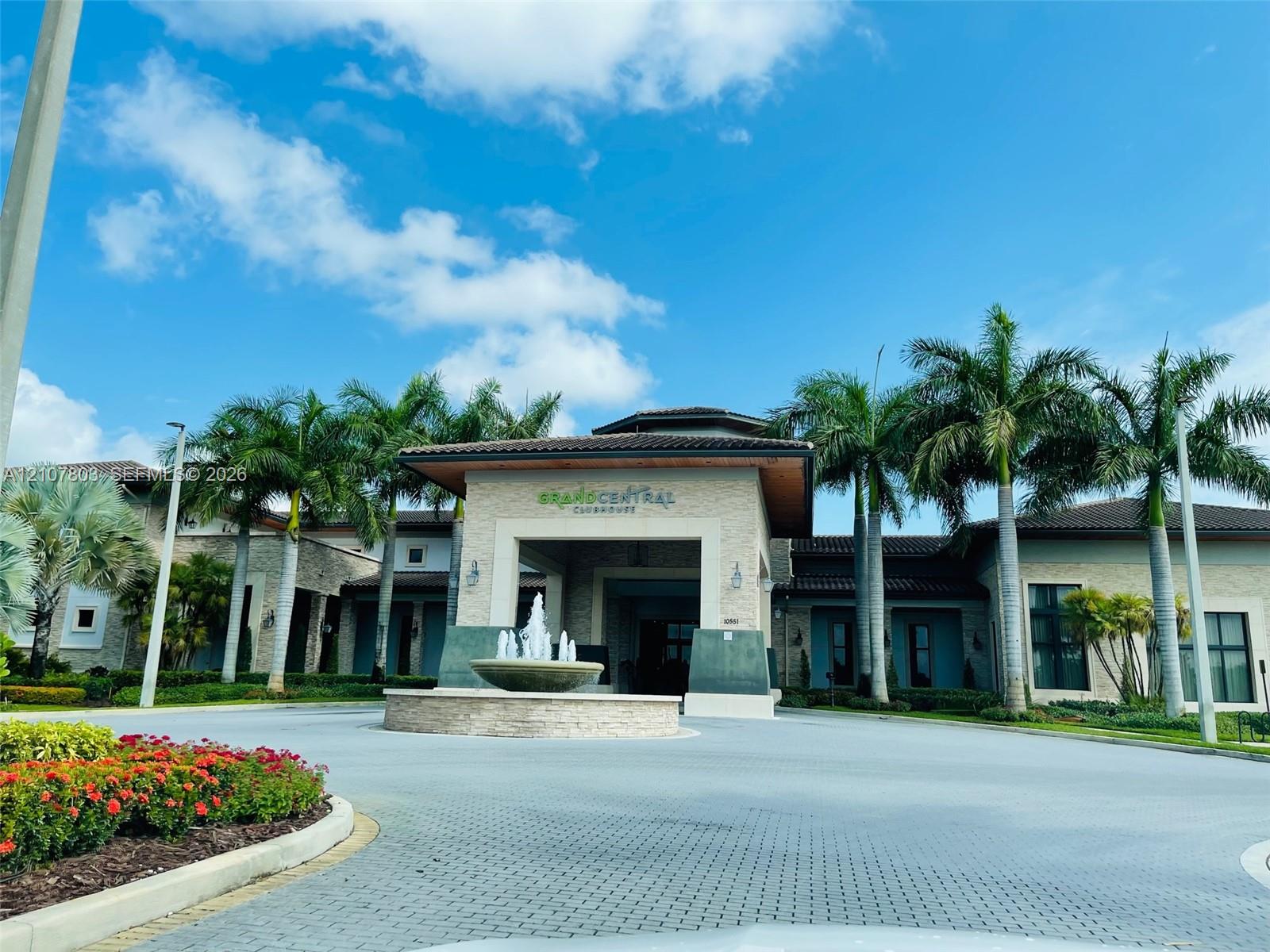 Clubhouse exterior featuring a stone-clad facade, covered porte-cochre, decorative water fountain, and mature palm trees