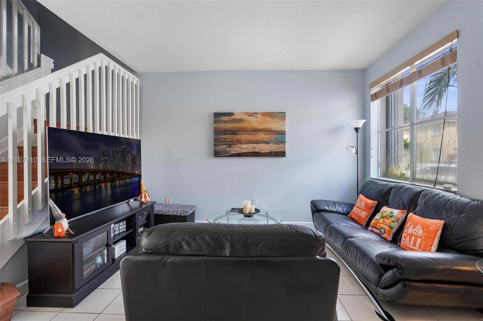 Living area featuring ceramic tile flooring, a large window with blind, and a white staircase with wooden handrail