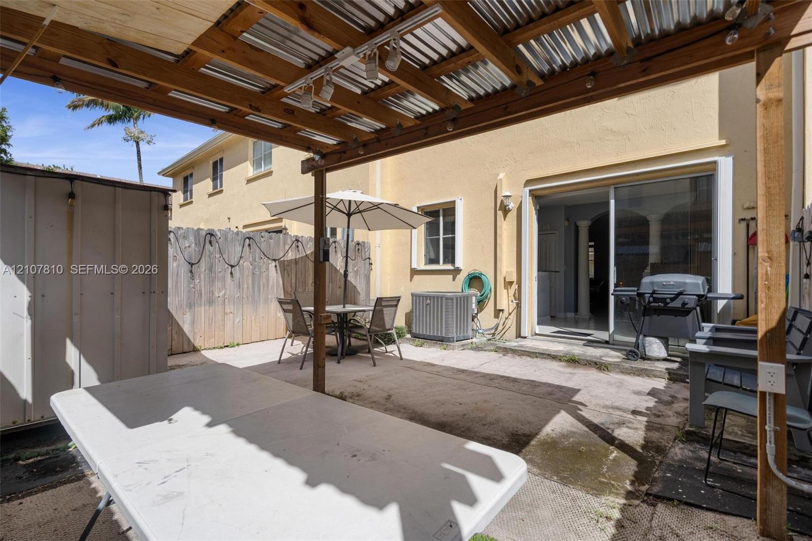 Paved rear patio featuring a corrugated metal and wood-beam pergola, wood privacy fencing, and sliding glass door access. Outdoor furniture, BBQ Grill and Outside TV stays.