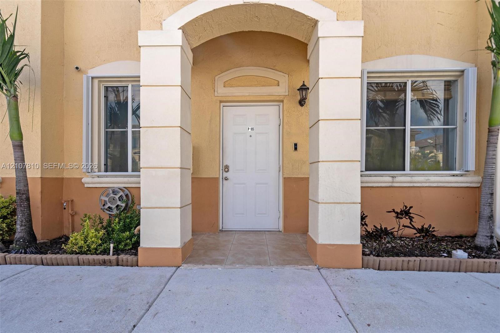 Stucco exterior entrance featuring a covered arched portico with decorative columns, tile porch flooring, and two dual-pane windows with integrated shutters Stucco exterior entrance featuring a covered arched portico with decorative columns, tile porch flooring, and two dual-pane windows with integrated shutters