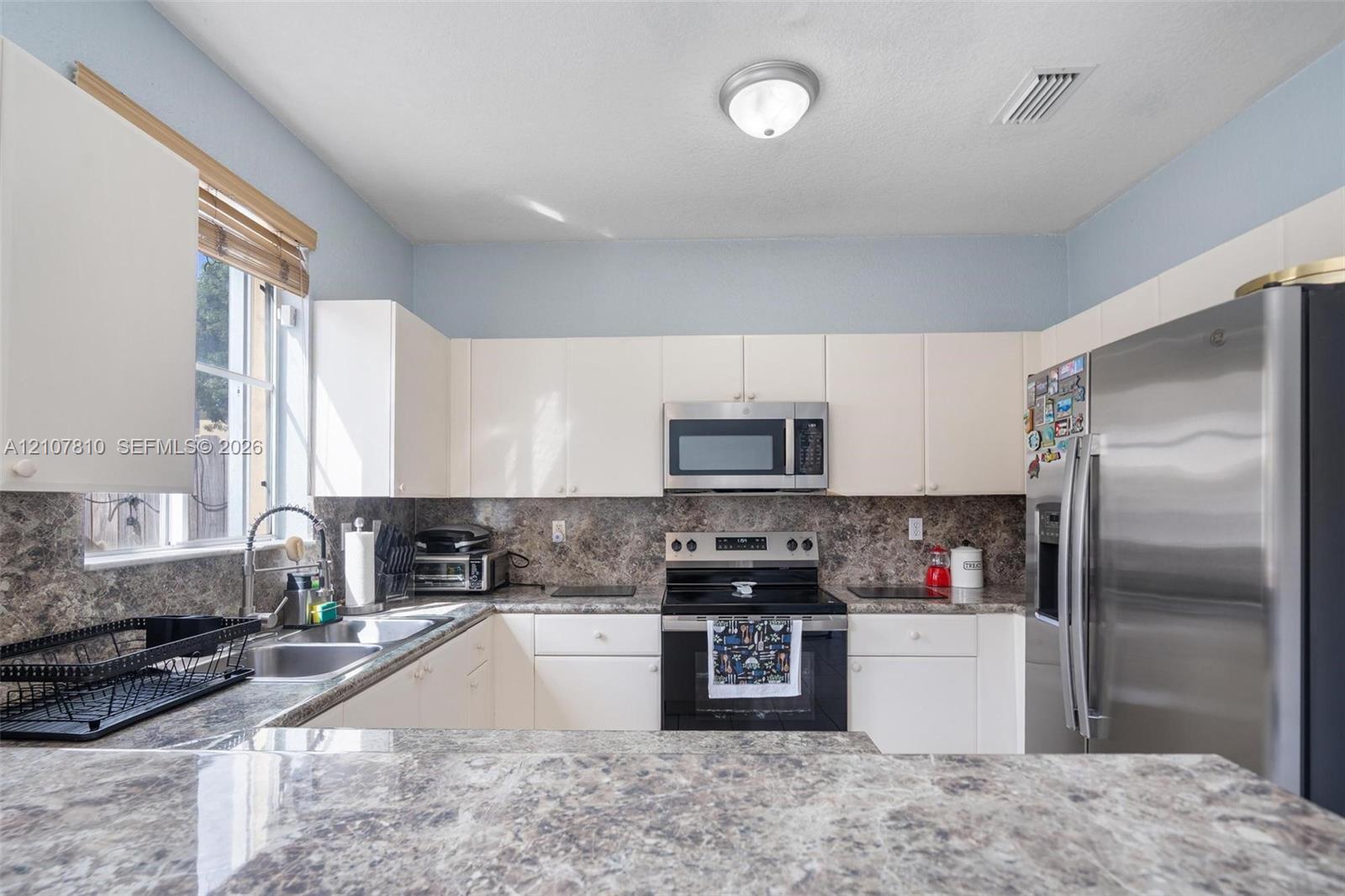 Kitchen featuring white cabinetry, stone-patterned countertops, stainless steel refrigerator, over-the-range microwave, and dual-basin sink with a gooseneck faucet