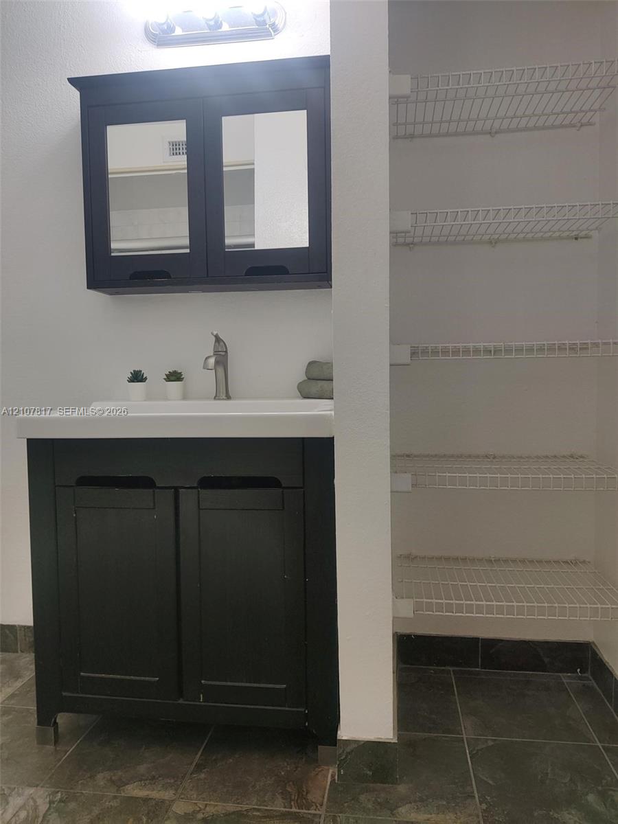 Dark-finish vanity with integrated rectangular sink and matching mirrored cabinet, accompanied by a multi-level wire shelving unit and tile flooring