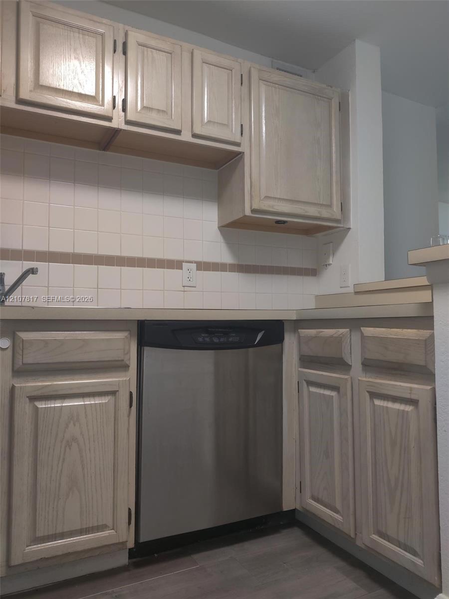 Kitchen featuring light-finish wood cabinetry, stainless steel dishwasher, square tile backsplash with decorative accent inlay, and wood-finish flooring