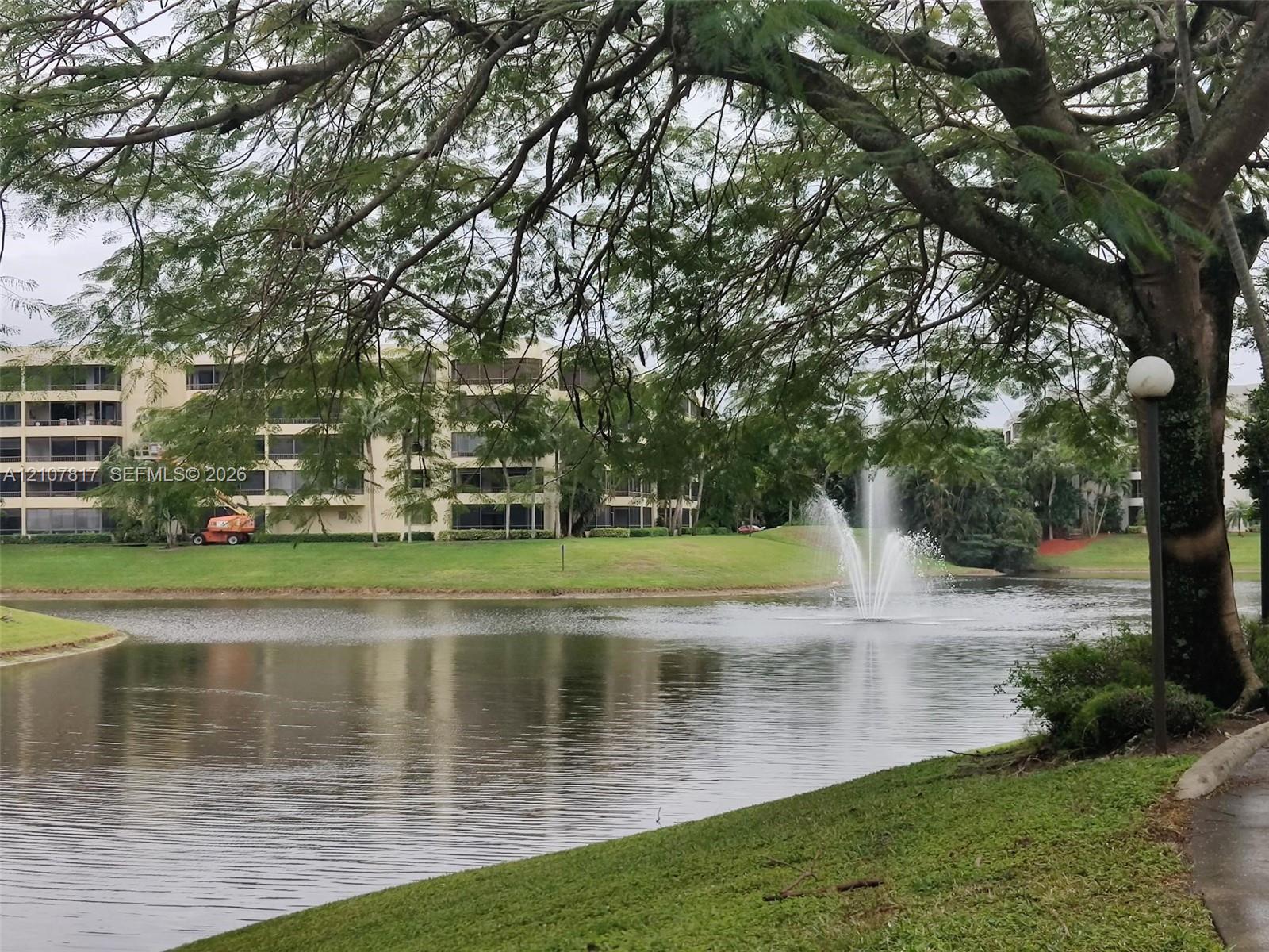 Waterfront setting featuring a central fountain, manicured lawn, mature trees, and paved walkway