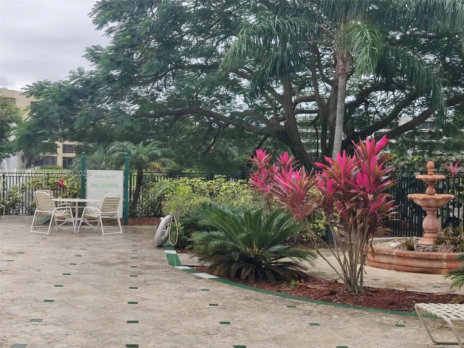 Outdoor patio featuring stone-tile flooring with decorative inlays, a tiered stone fountain, mature tropical landscaping, and metal perimeter fencing