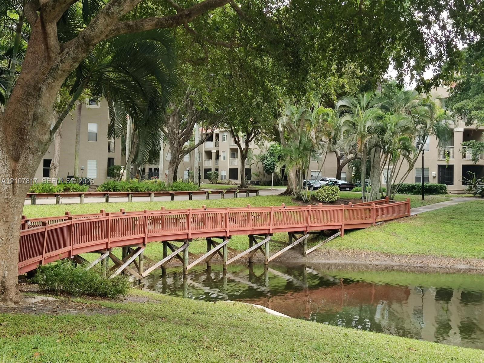 Pedestrian bridge with wooden railing over a landscaped pond, surrounded by mature trees and manicured green space