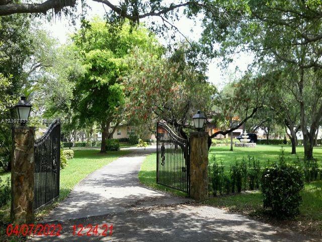 Gated entrance featuring stone masonry pillars, wrought iron fencing, mounted exterior lighting fixtures, and a landscaped grounds area with mature trees and a fountain