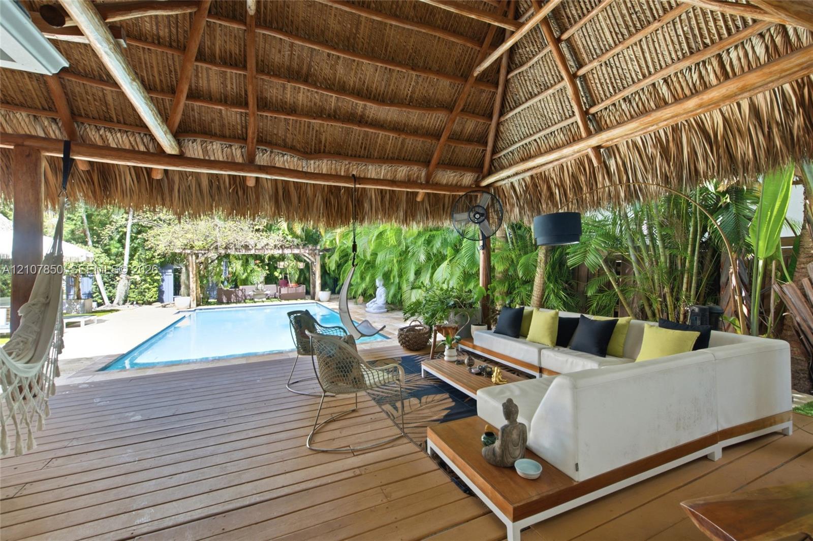 Open-air pavilion featuring a thatched roof, wood-finish decking, and a built-in wall-mounted fan, overlooking an in-ground swimming pool