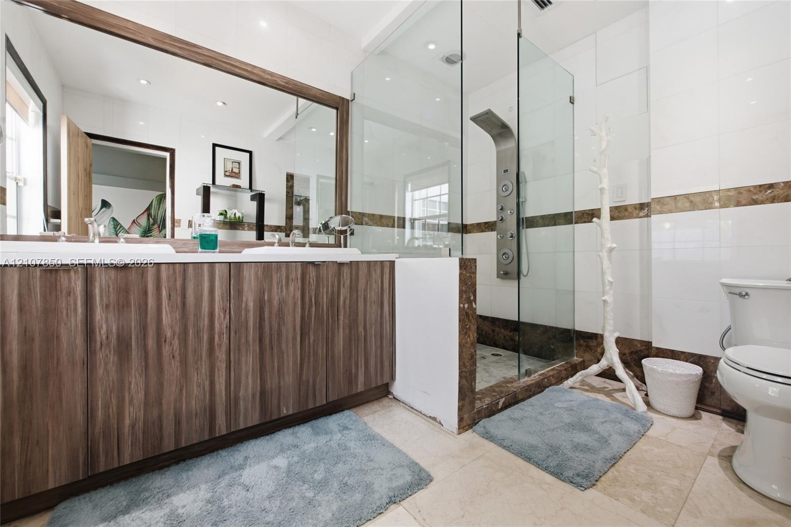Dual-sink vanity with wood-finish cabinetry, full-width mirror, walk-in shower with frameless glass enclosure, and stone-tile flooring